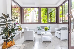 A bright, sunlit room with white wicker furniture, large windows, and a potted plant. - Hotel Luana  (Riccione)