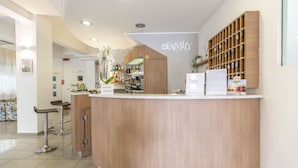 A reception area with a wooden counter, bar stools, a wall-mounted display, and a kitchen area in the background. - Hotel Luana  (Riccione)