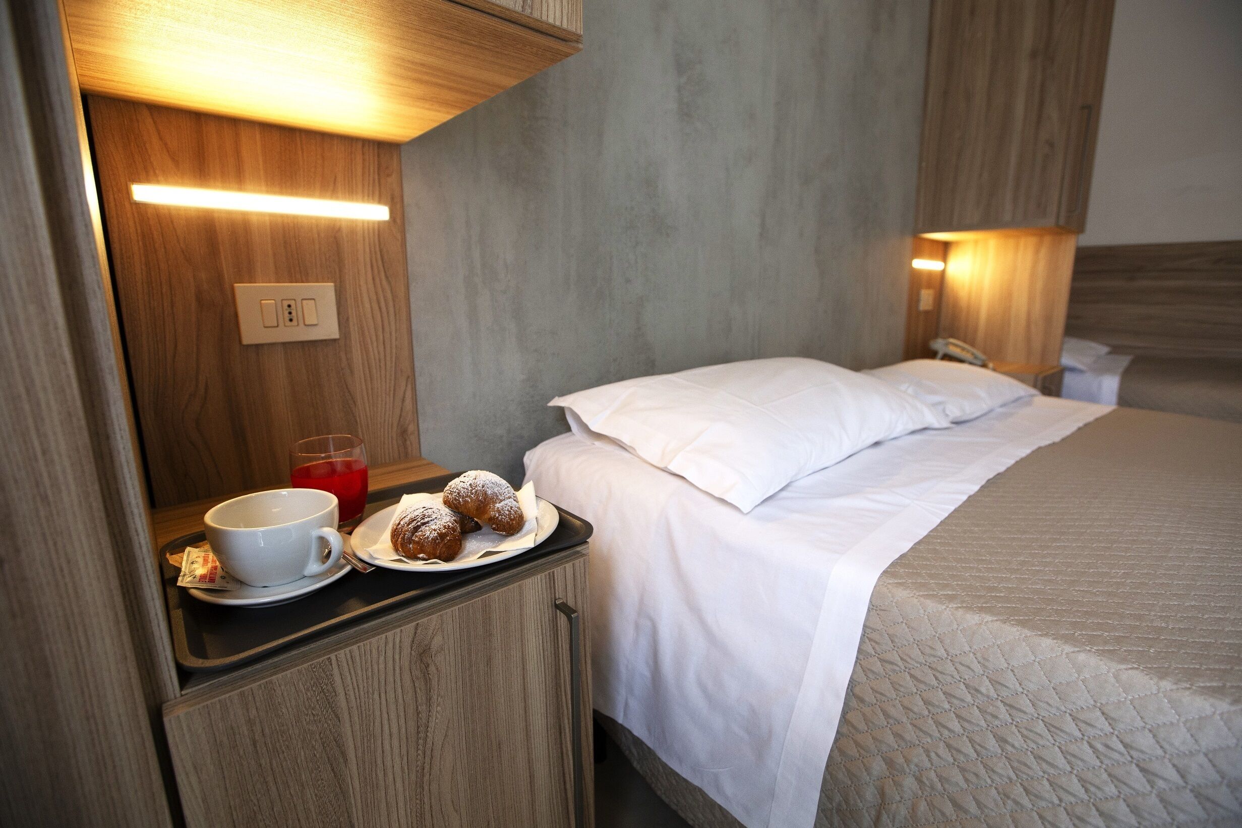 Habitación de hotel con cama, mesita de noche y una pequeña mesa con taza, plato y pasteles.