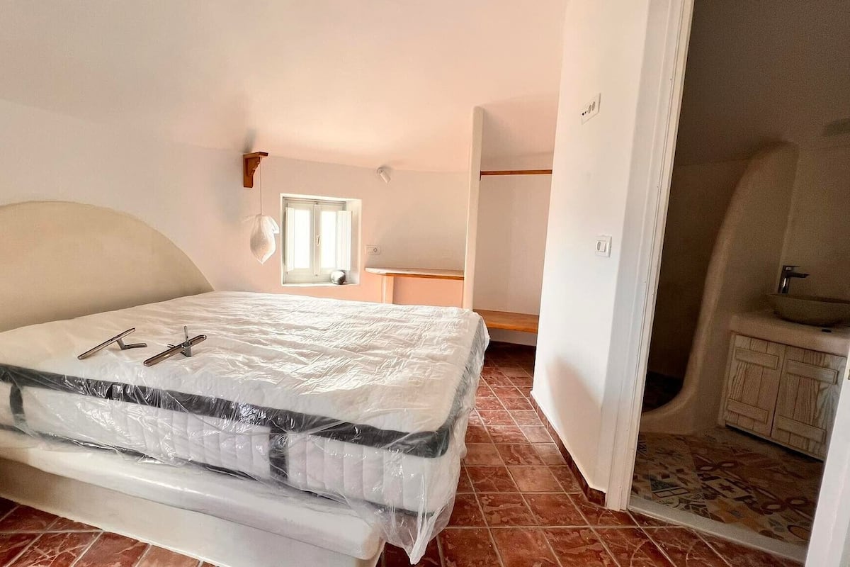 Un dormitorio con cama, mesita de noche, ventana y un baño visible a través de una puerta abierta.