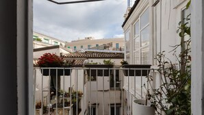 Un balcón estrecho con barandilla de metal, plantas en macetas y vistas a edificios cercanos.