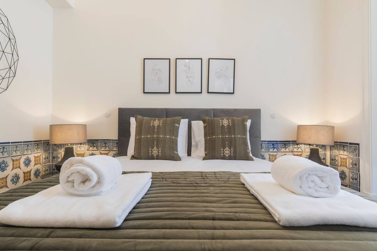 A hotel room with a bed, two pillows, a headboard, a wall with three framed pictures, and a lamp on either side of the bed.