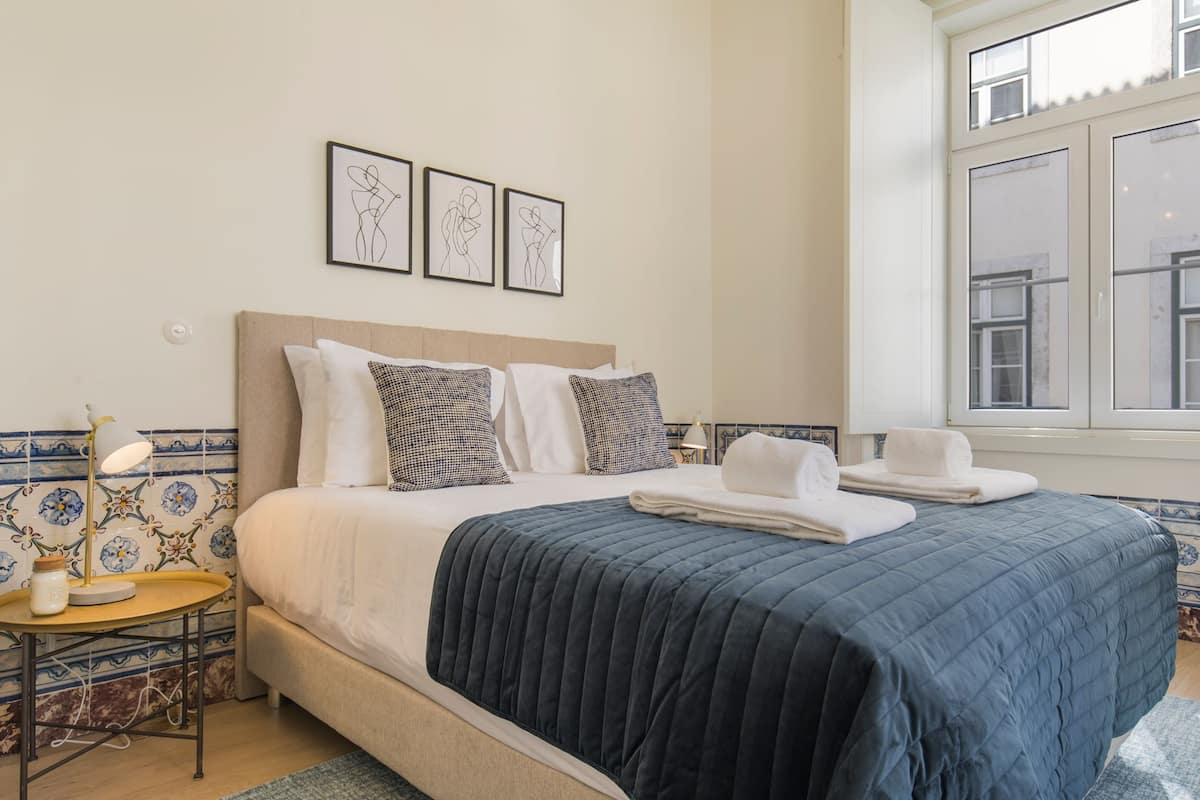 A neatly made bed with a dark blue quilt, a bedside table with a lamp, and a window with a view of buildings.
