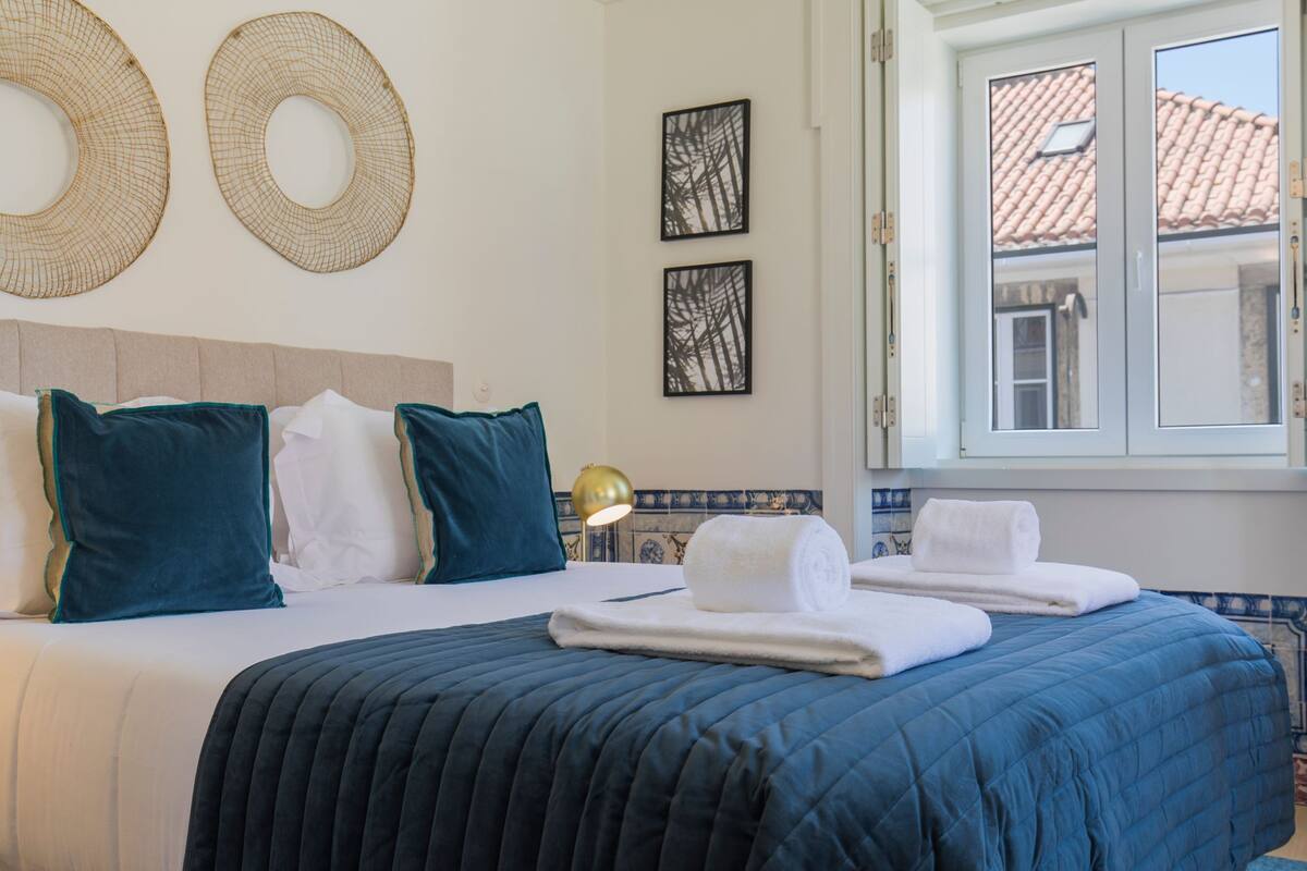 A hotel room with a bed, two wall-mounted circular decorations, a window with a view of a tiled roof, and a lamp on a bedside table.
