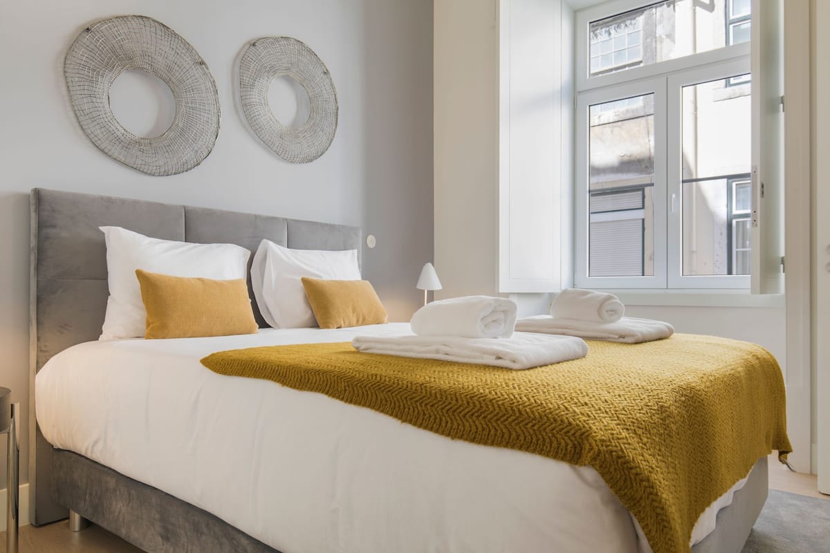 A neatly made bed with white and mustard bedding, two decorative wall pieces, and a window with a view of the outside.