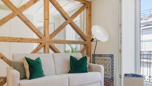 Habitació amb un sofà blanc, coixins verds, una estructura de sostre de fusta, una làmpada blanca i vista cap a l'exterior.