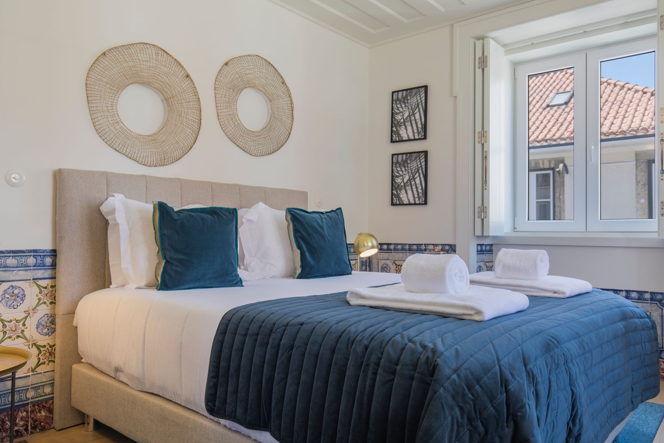 A neatly made bed with a blue quilt, white pillows, and a beige headboard. There are two wall decor pieces above the bed and a window with a view of a building.