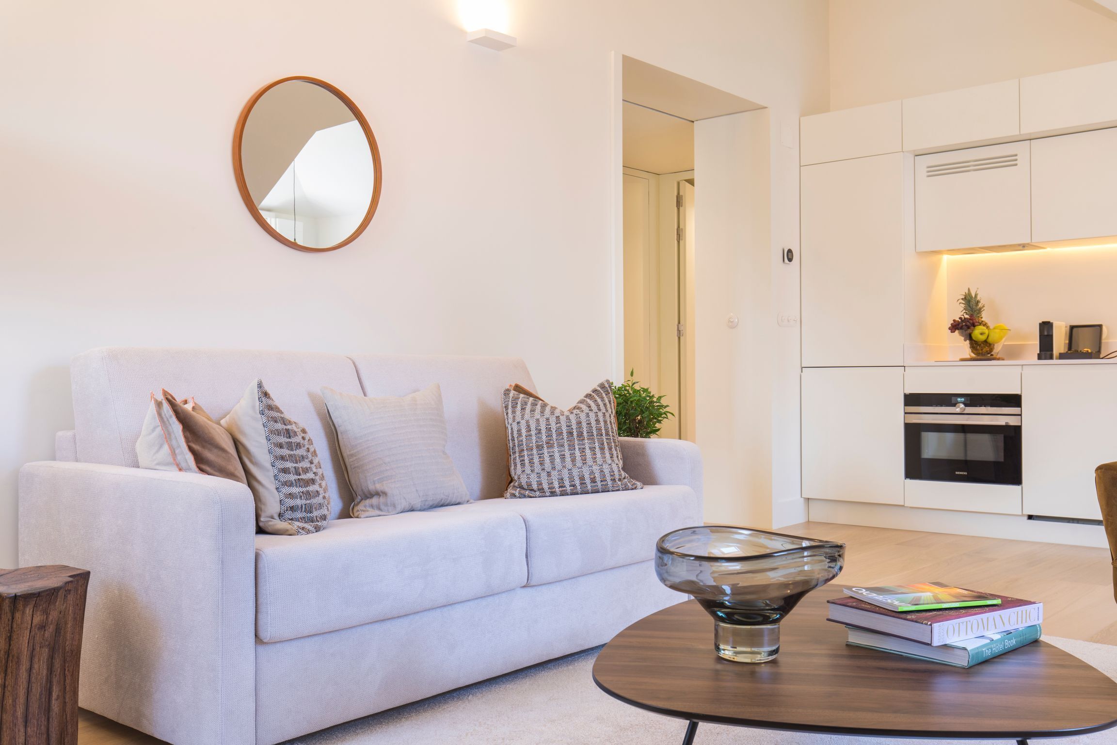 Ein modernes Wohnzimmer mit einem hellen Sofa, einem Glastisch und einer Küche im Hintergrund.