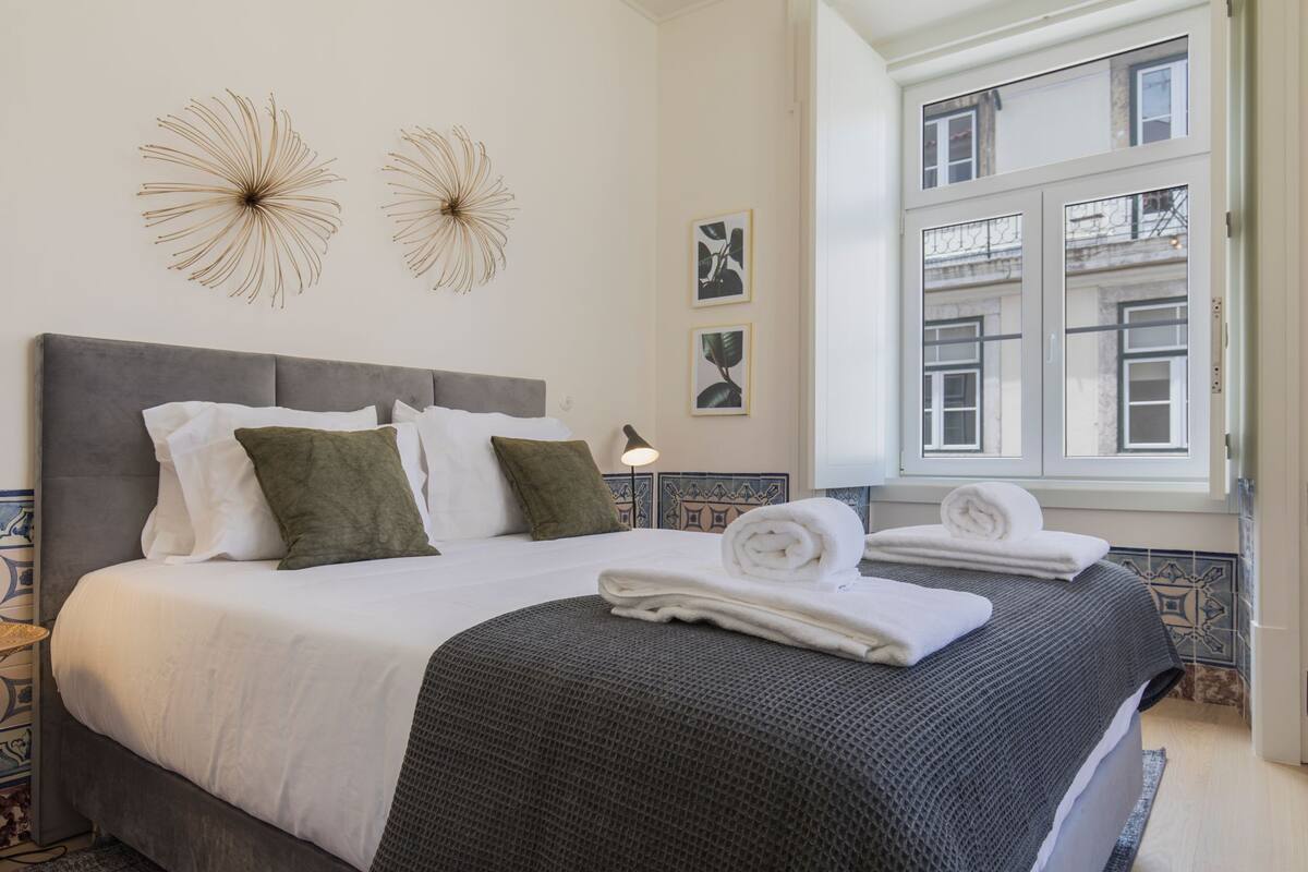 A neatly made bed with white and green pillows, a grey headboard, and a grey bedspread. There are two wall art pieces above the bed and a window with a view of buildings.