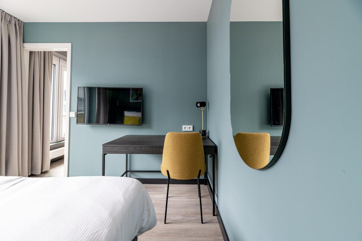 A hotel room with a bed, a desk, a chair, a flat-screen TV, and a mirror.