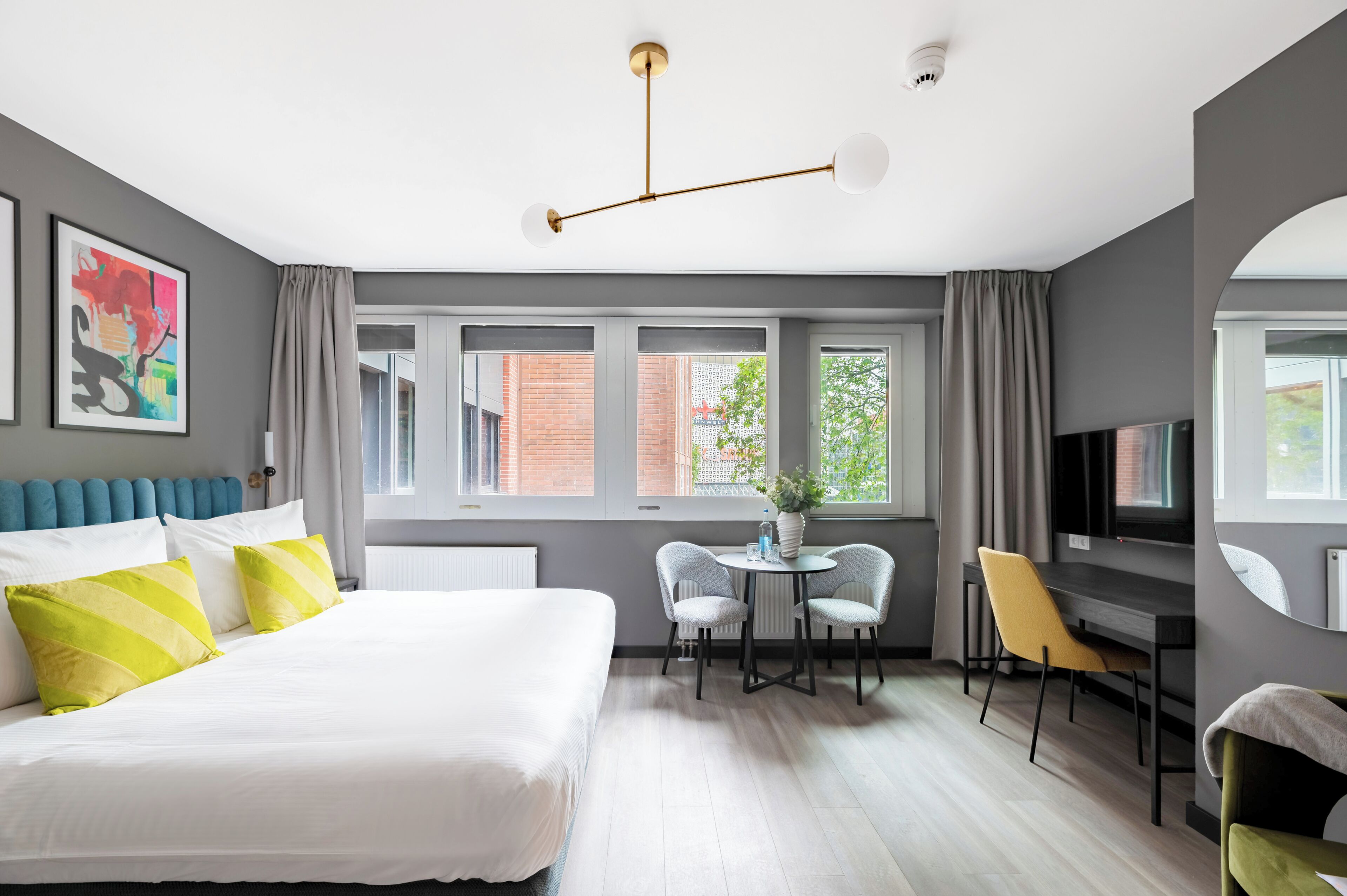 A modern hotel room with a large bed, a desk with two chairs, a television, and a window with a view of a brick building.