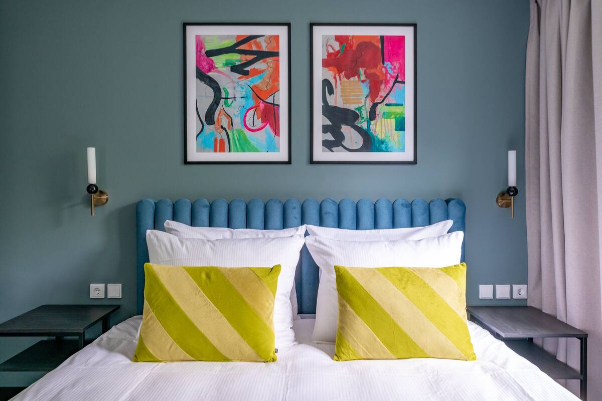 A hotel room with a blue headboard, two abstract paintings on the wall, and a bed with white and yellow striped pillows.