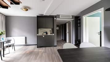 A modern hotel room with a dark wood dining table, a kitchenette with a built-in oven, a radiator, and a large window.