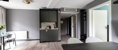 A modern hotel room with a dark wood dining table, a kitchenette with a built-in oven, a radiator, and a large window.