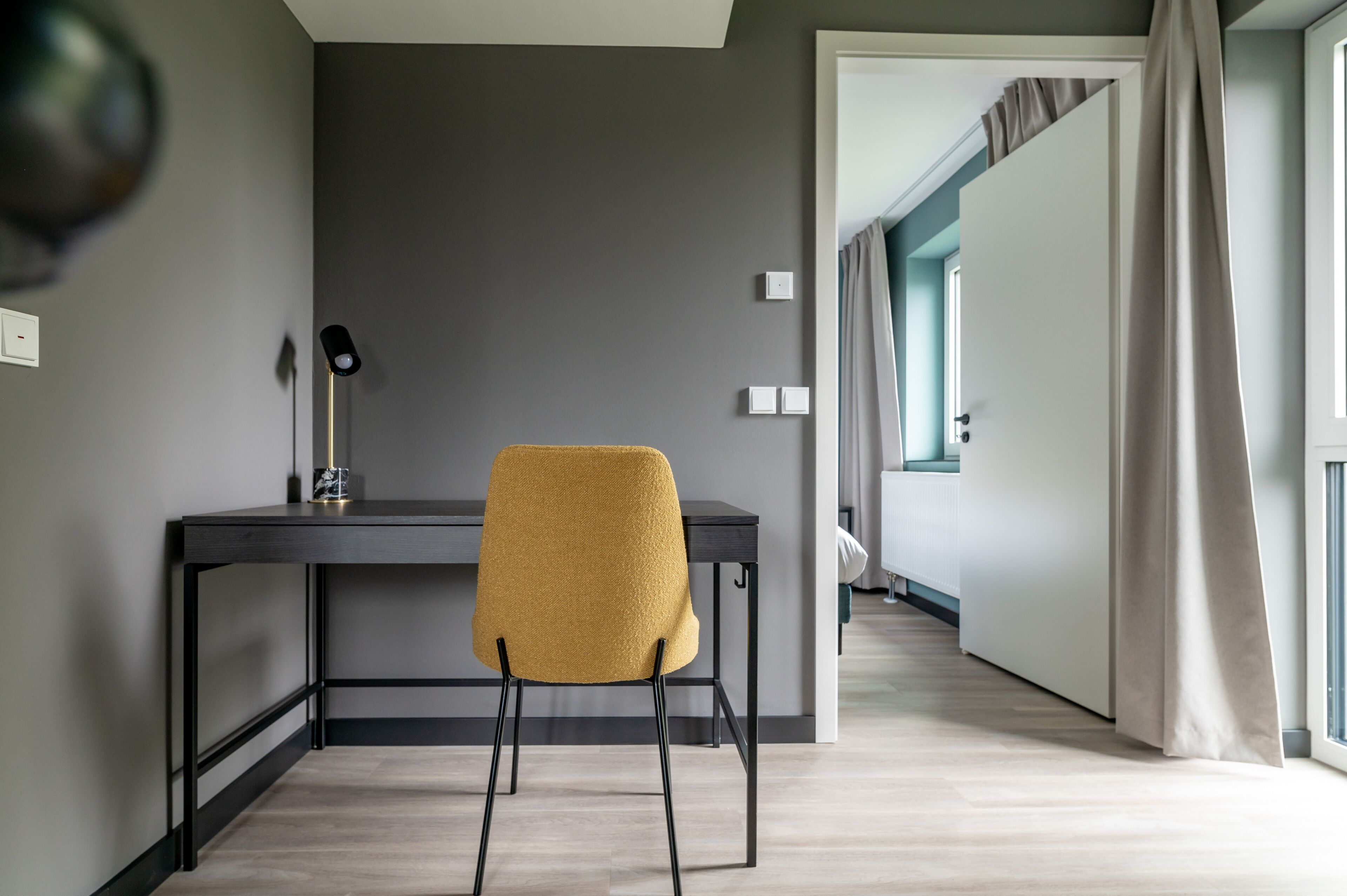 Quarto moderno com parede cinza escura, piso de madeira, escrivaninha preta e cadeira amarela, além de uma janela com cortinas.
