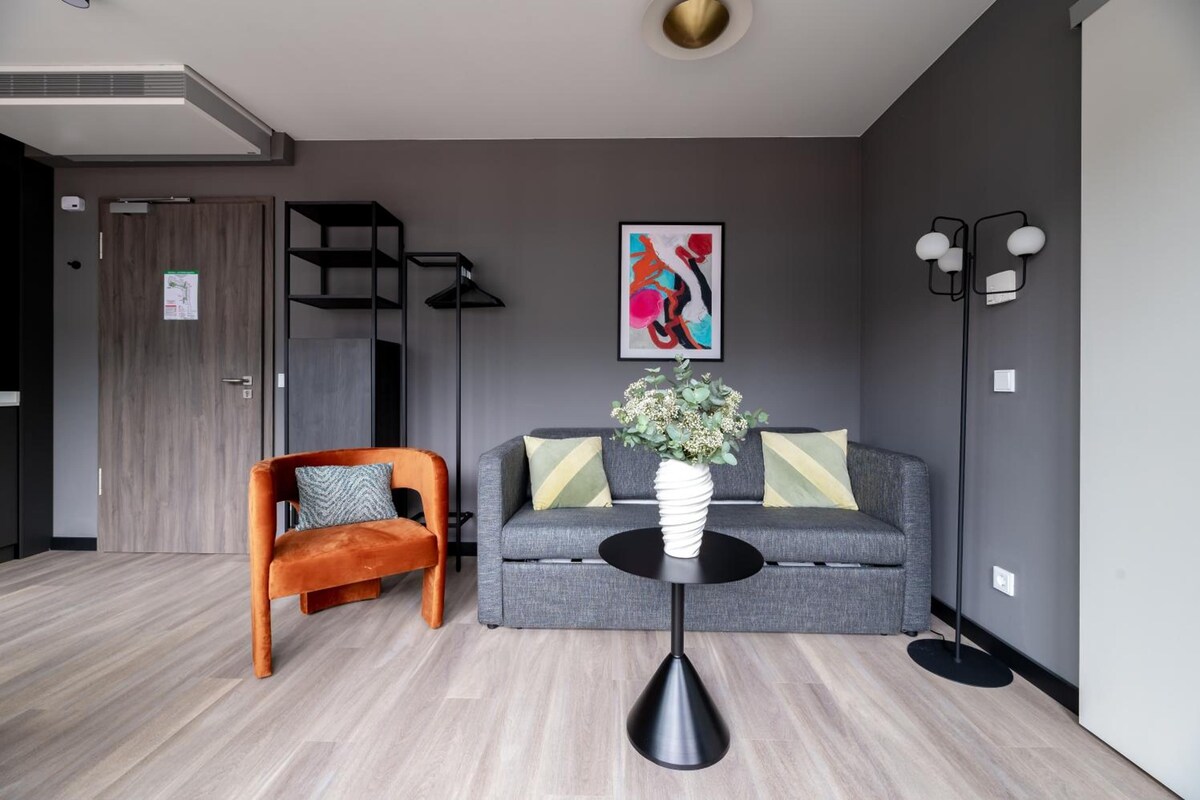 A modern living room with a grey sofa, an orange chair, a black floor lamp, and a round table with a vase of flowers.