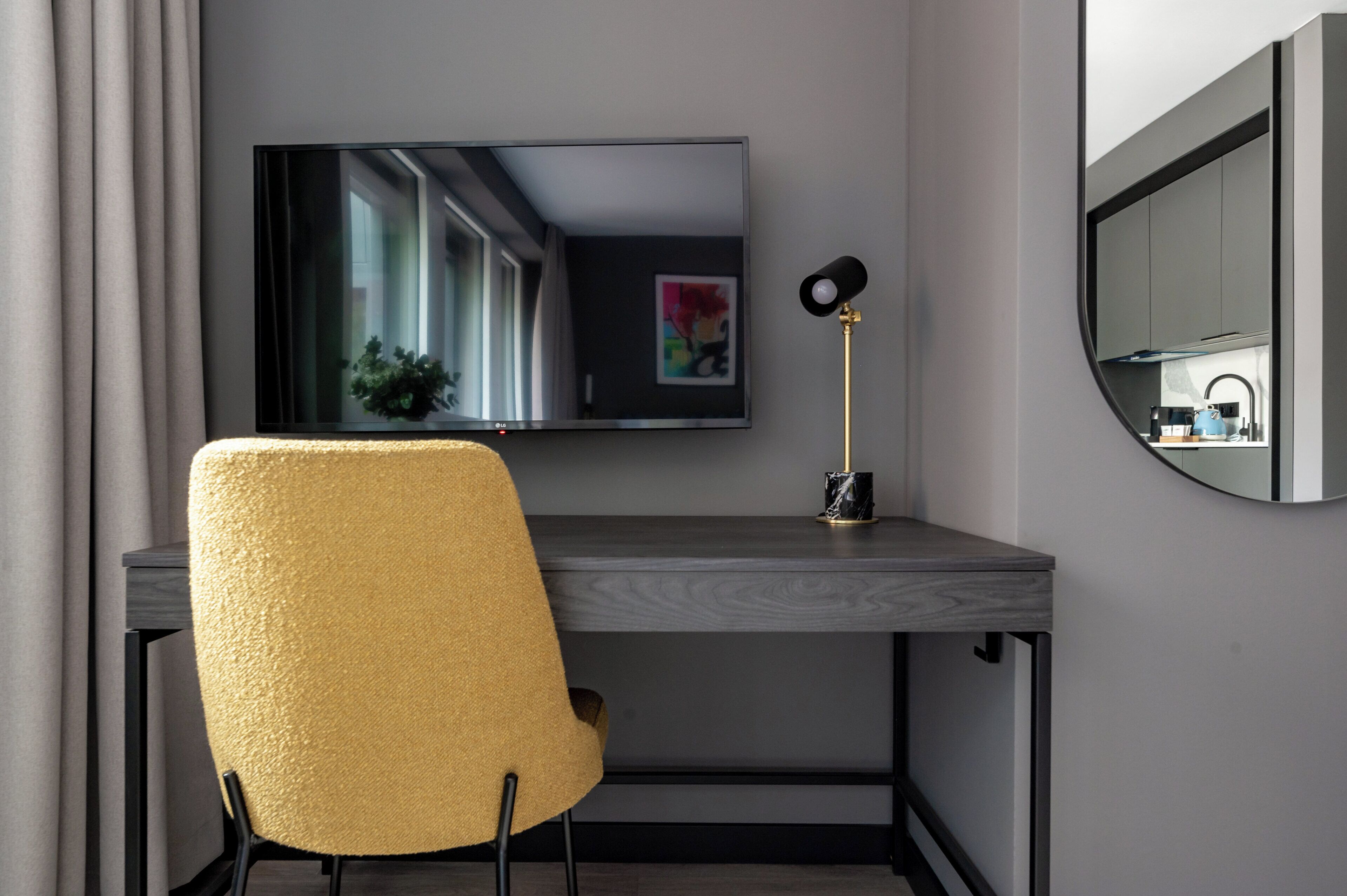 Ein modernes Zimmer mit Schreibtisch, Stuhl, Fernseher, Lampe und Blick auf eine Küche.