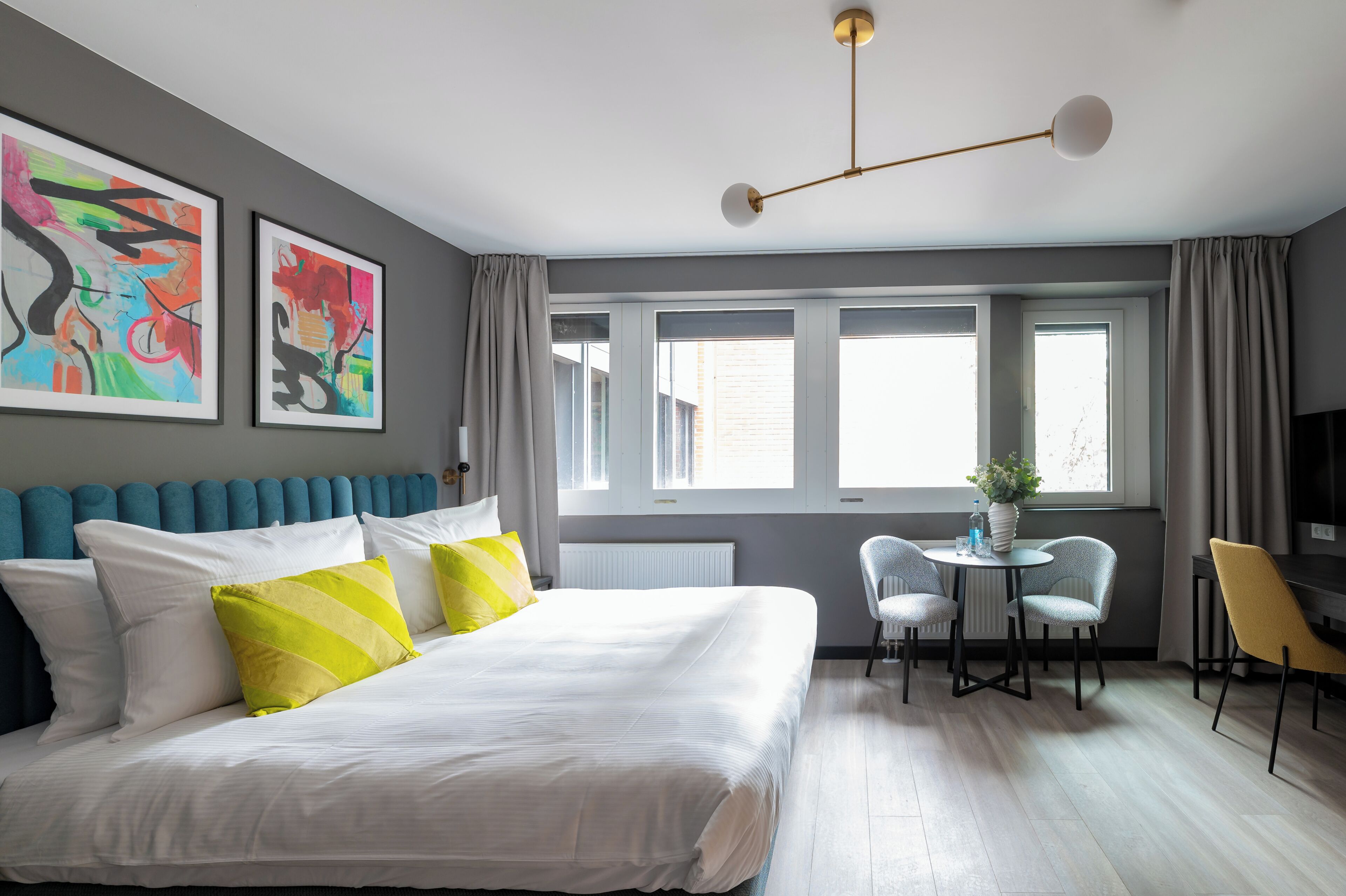 Un dormitorio moderno con una cama grande, un escritorio con dos sillas, un ventanal con cortinas y arte abstracto en la pared.