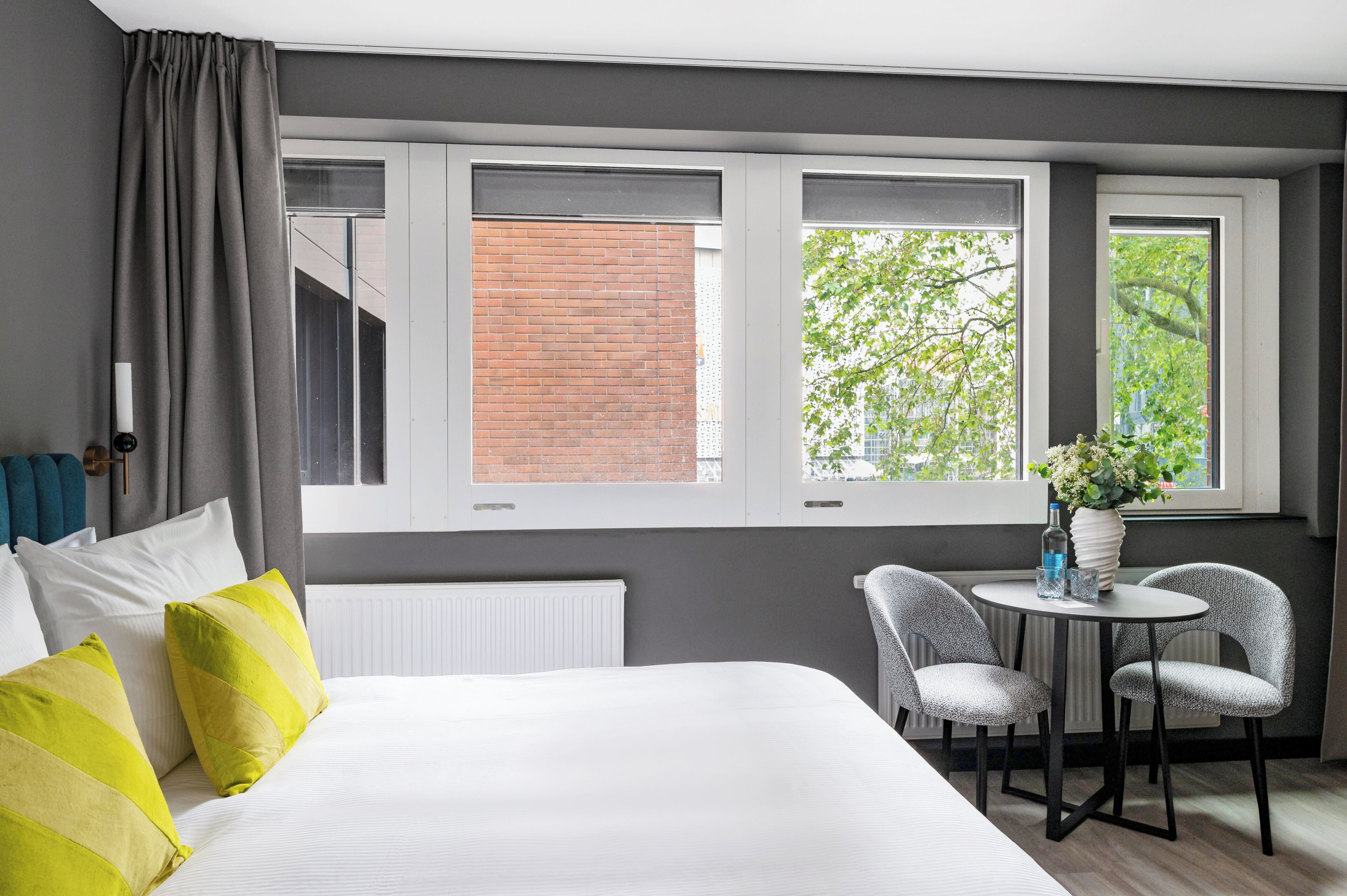 Una habitación de hotel con una cama, dos sillas, una mesita y tres ventanas.