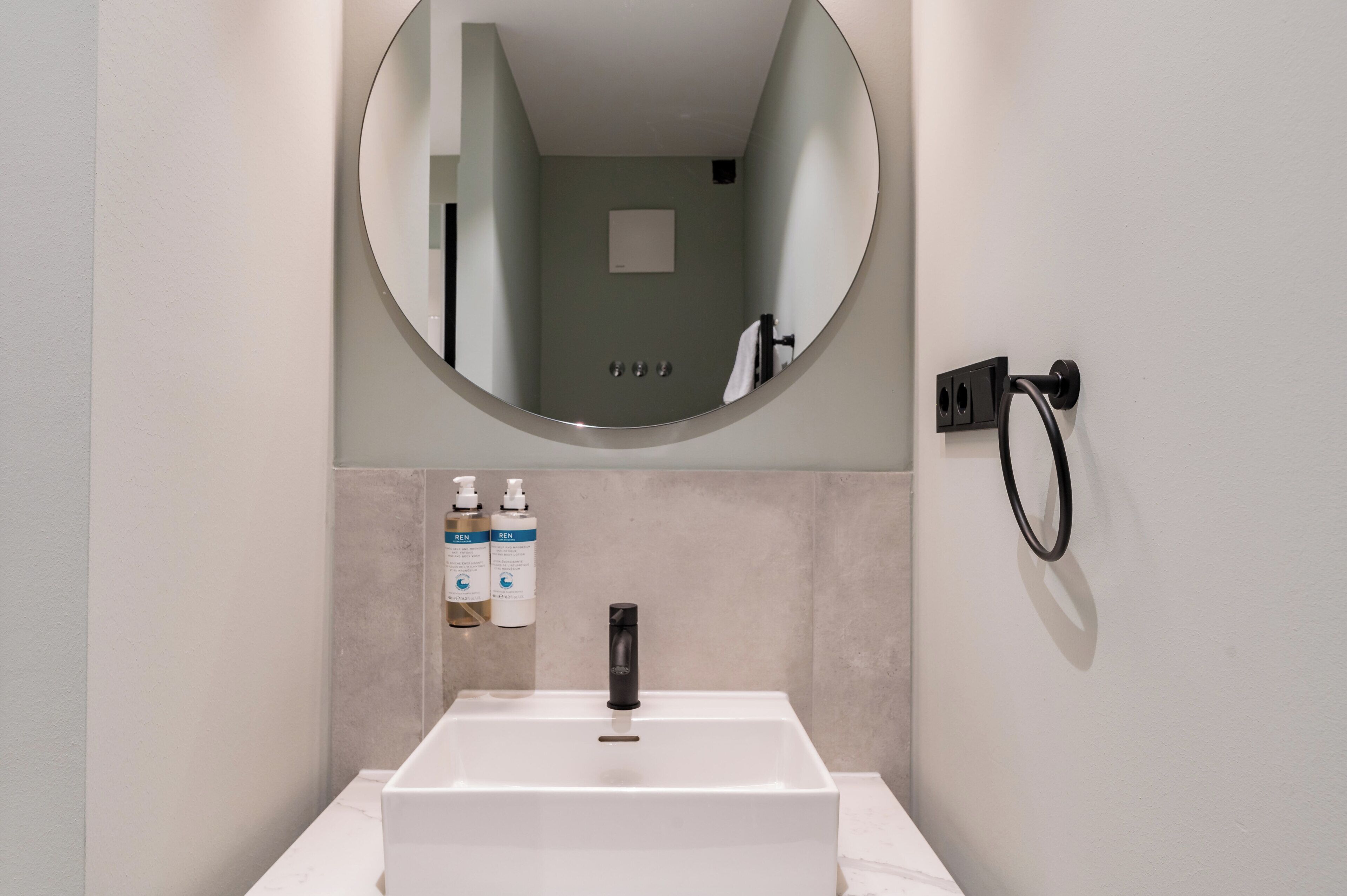 Une salle de bain avec un lavabo blanc, un miroir rond et deux flacons de savon pour les mains.