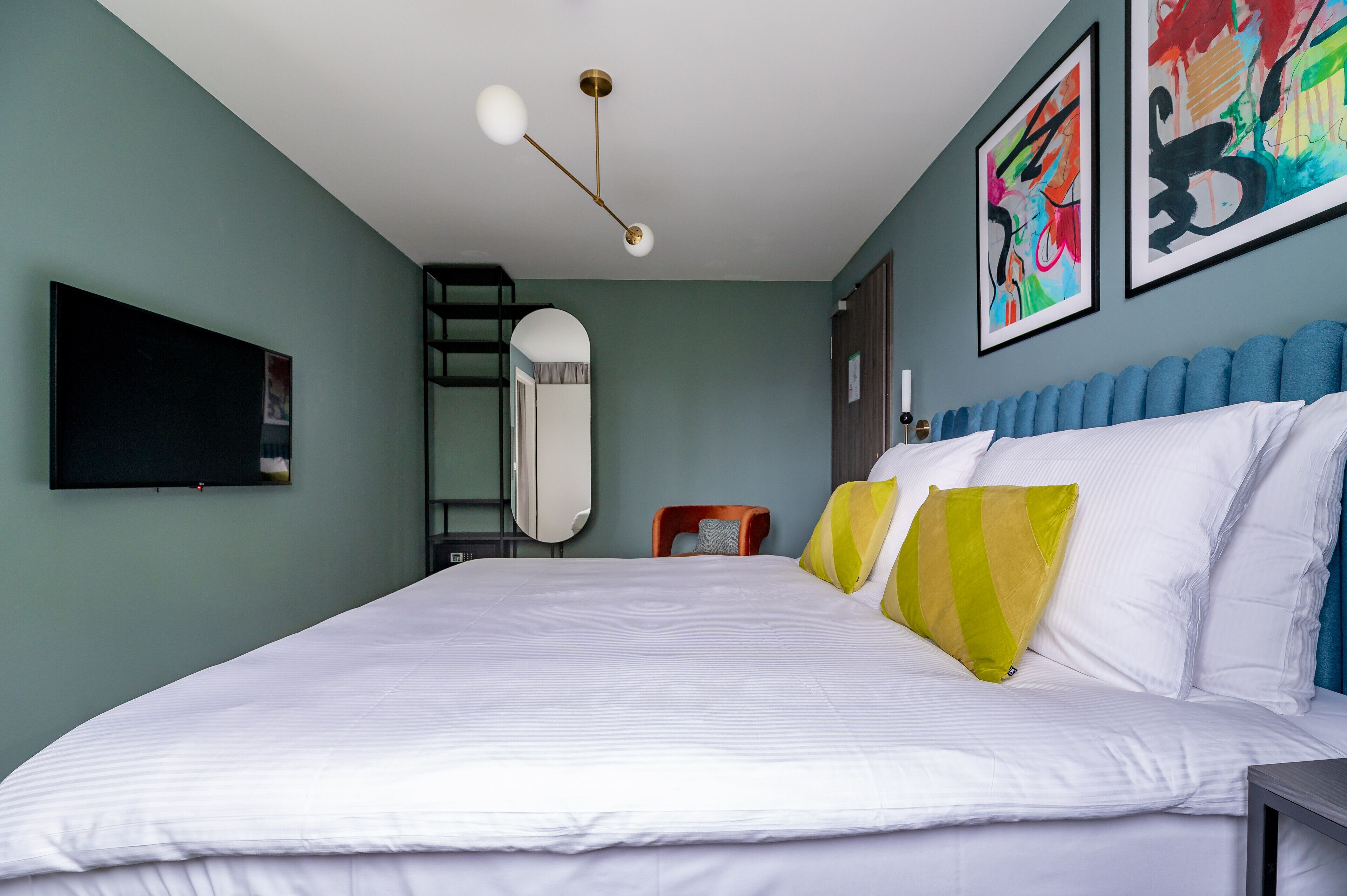 A neatly made bed with white linens, a modern ceiling light fixture, and colorful abstract art on the walls.