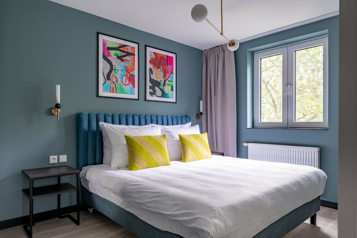 A bedroom with a bed, two framed abstract paintings, a bedside table, and a window with curtains.