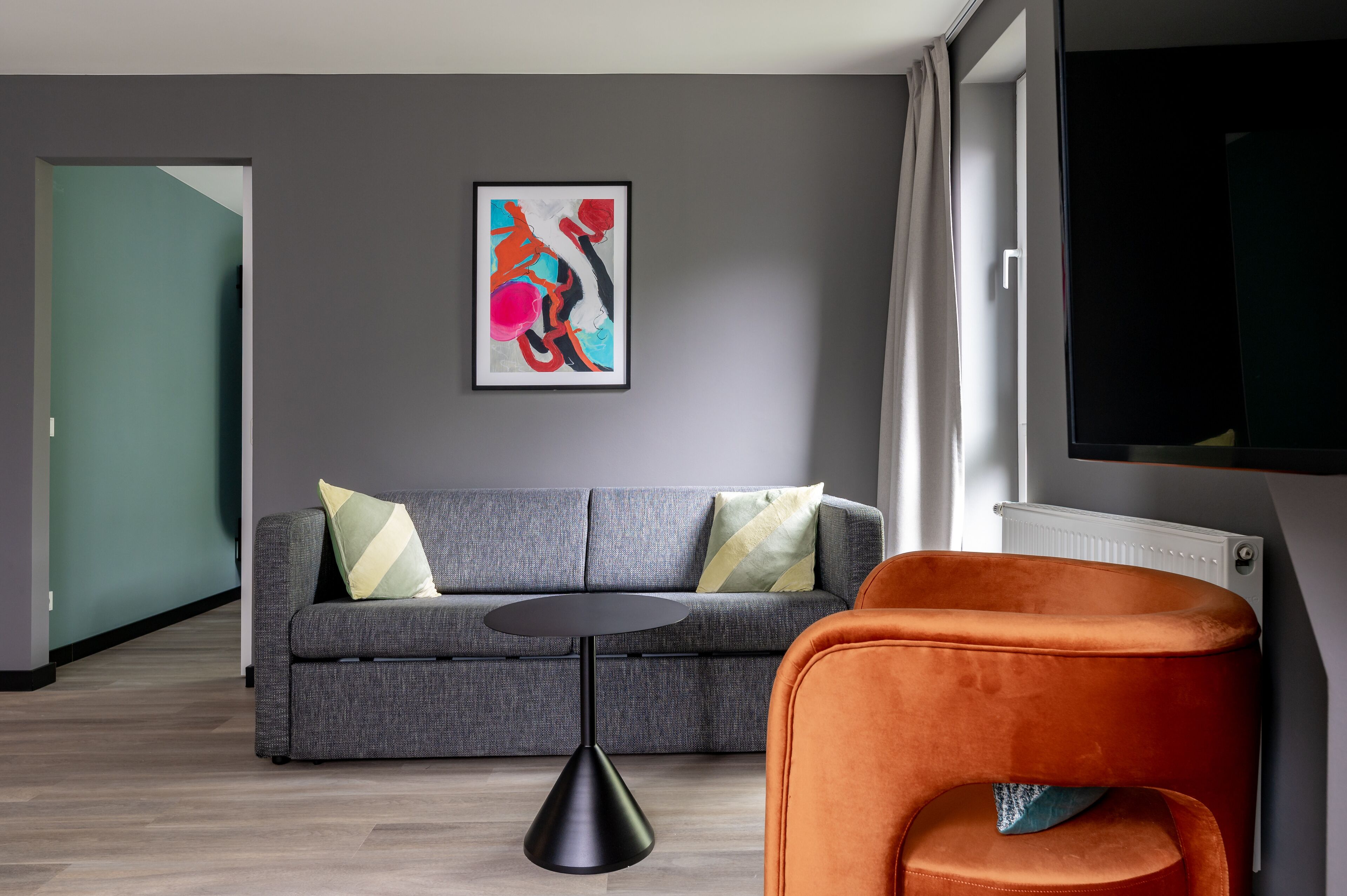 A modern living room with a grey sofa, an orange armchair, a black coffee table, and a colorful abstract painting on the wall.