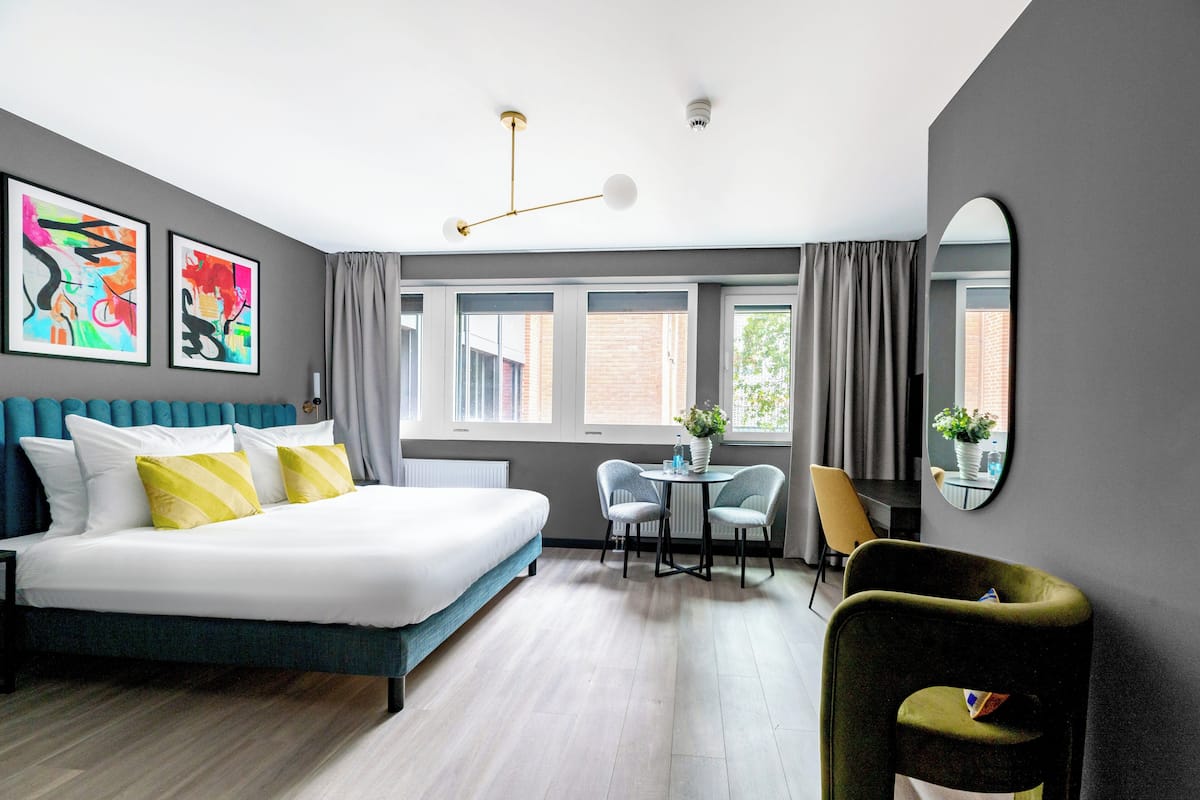 A modern hotel room with a bed, a dining table, two chairs, a green armchair, and artwork on the walls.