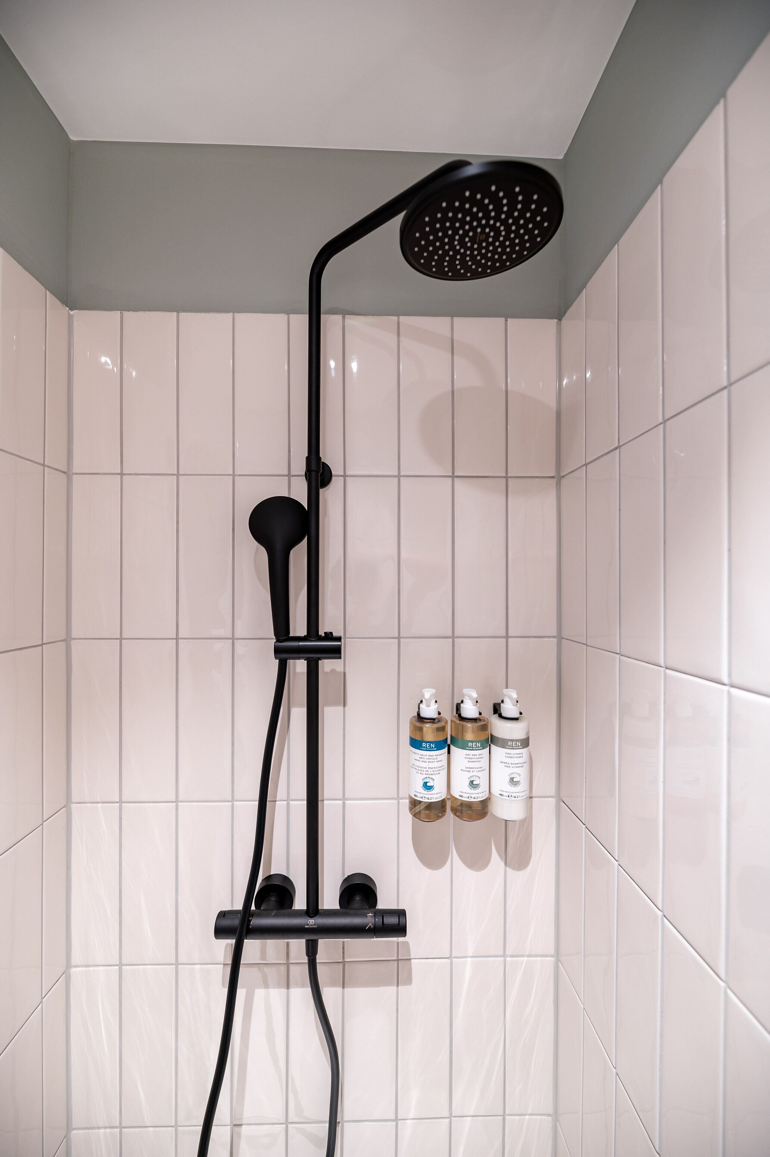 Eine Dusche mit einem schwarzen Duschhandstück, einer an der Wand montierten Bedienkonsole und drei Flaschen Flüssigseife auf einem Regal.