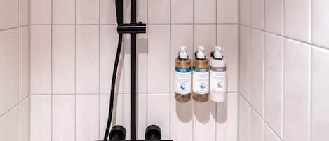 A shower with a black handheld showerhead, a wall-mounted control panel, and three bottles of liquid soap on a shelf.