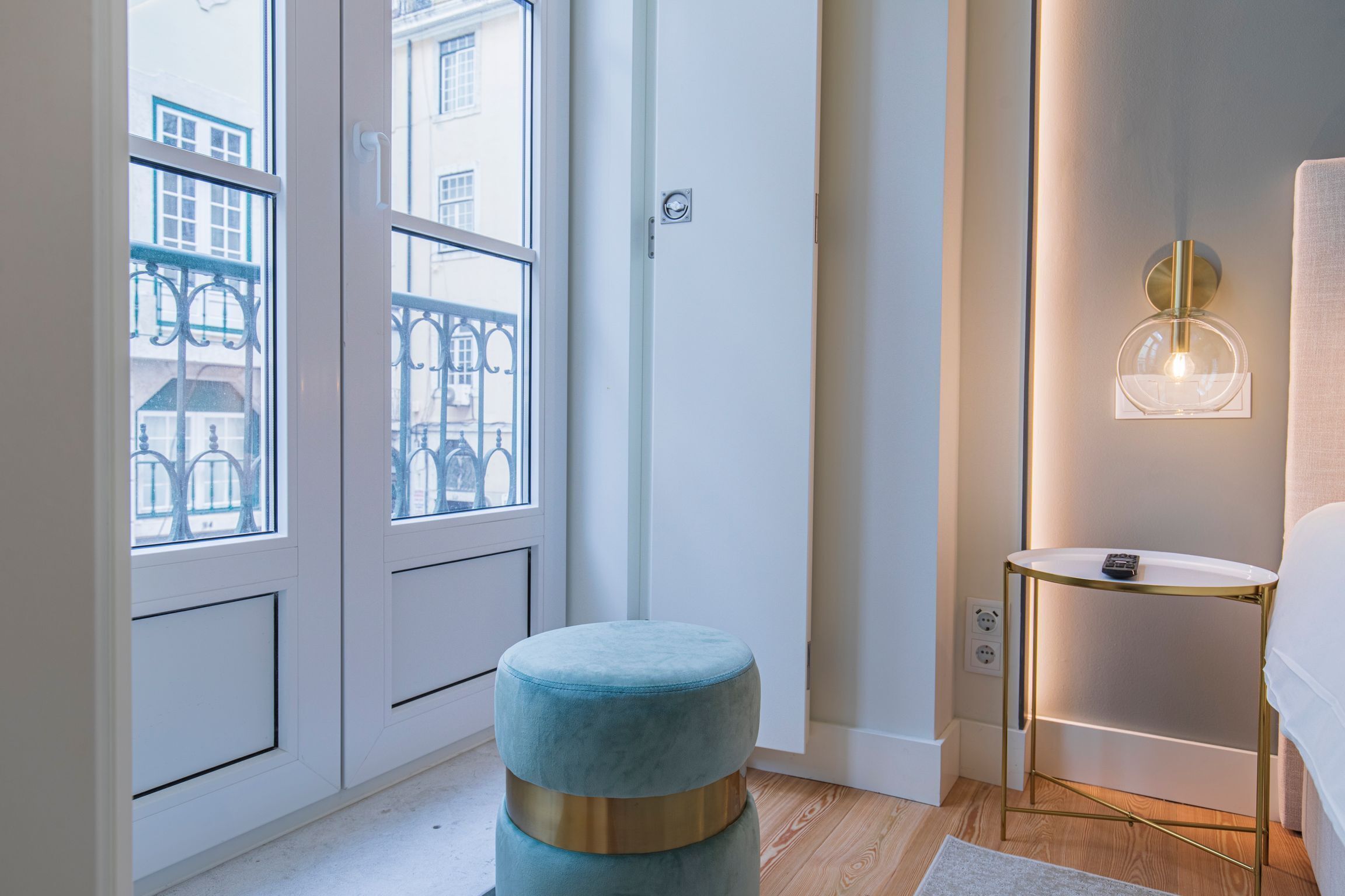 A modern bedroom with a large window, a bedside table with a lamp, and a round ottoman.