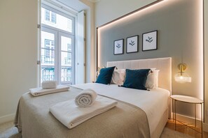 A neatly made bed with white linens and rolled towels, a bedside table with a lamp, and framed artwork on the wall. - Ando Living São Nicolau House (Lisbon)