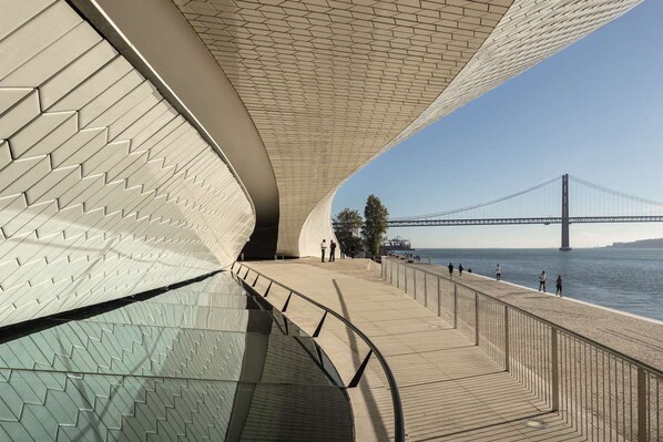 A waterfront promenade with a modern architectural design, featuring a curved walkway and a view of a bridge in the background. - Ando Living São Nicolau House (Lisbon)