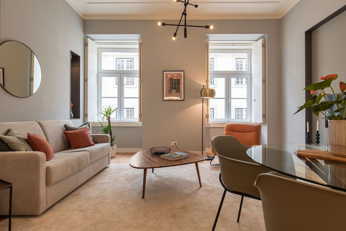Un salon moderne avec un canapé, une table basse et des fauteuils. On y trouve de grandes fenêtres, un miroir et un luminaire suspendu.