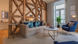 Uma sala de estar moderna com vigas de madeira expostas, um sofá com almofadas florais, uma mesa de centro redonda, uma poltrona azul e uma janela grande com vista.