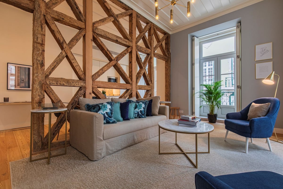 Un salon moderne avec des poutres apparentes, un canapé agrémenté de coussins à motifs floraux, une table basse ronde, un fauteuil bleu et une grande fenêtre offrant une vue dégagée.