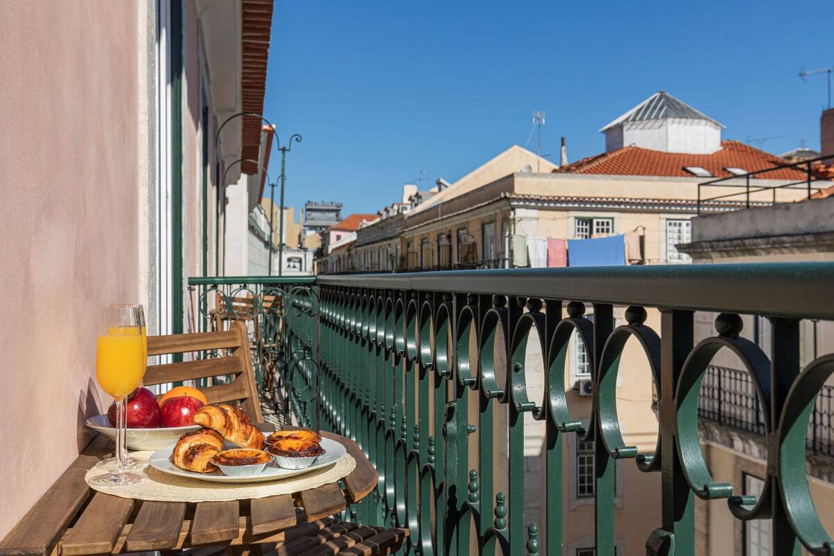 Un balcon avec une table préparée pour le petit-déjeuner, comprenant des croissants, des fruits et du jus d’orange.