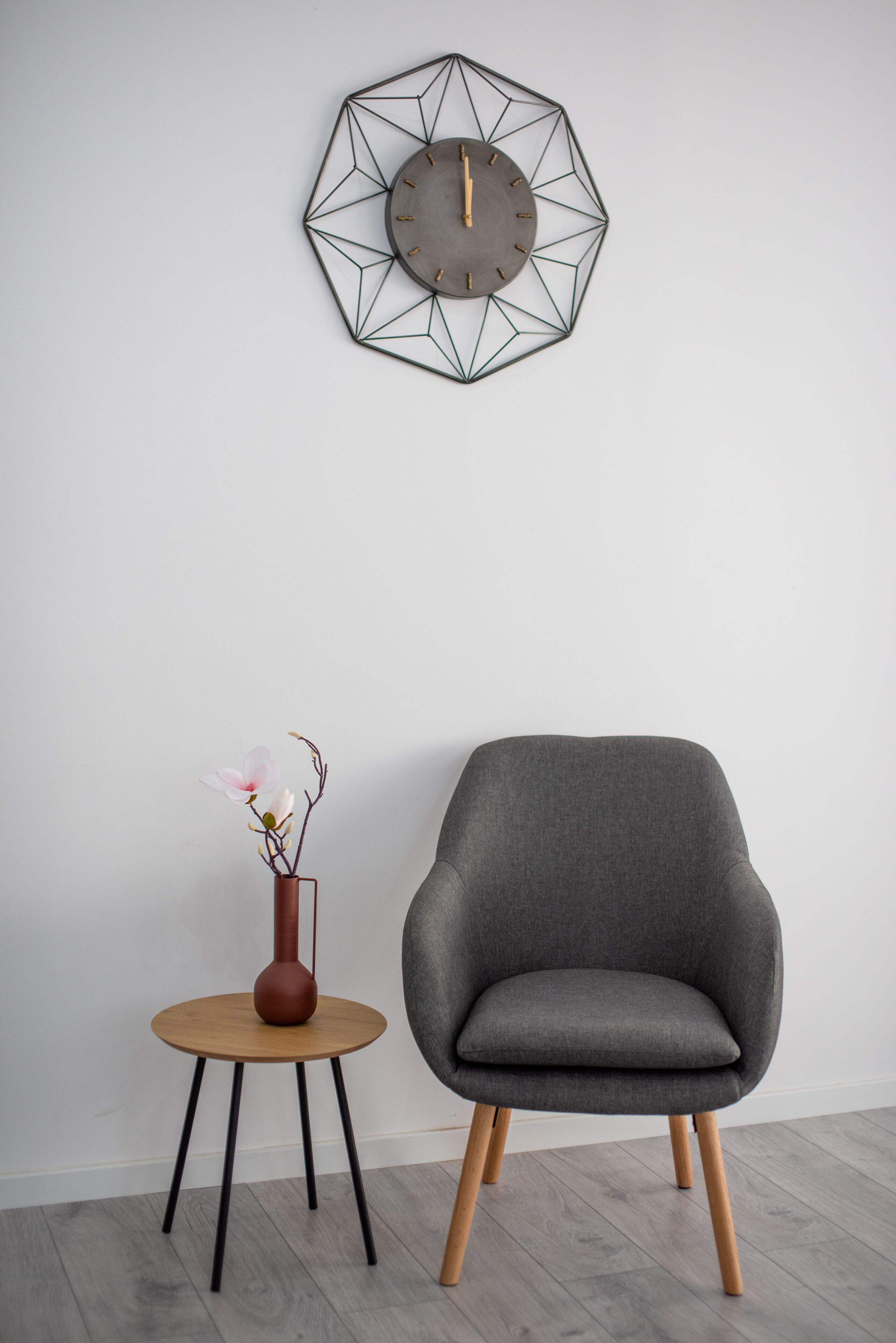 Een moderne kamer met een geometrische wandklok, een grijze fauteuil en een klein houten tafeltje met een bloemenvaas.