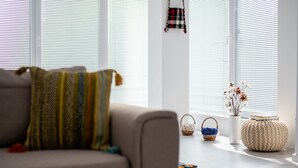 A living room with a sofa, patterned pillows, and a hanging bag. There are two baskets and a vase with dried flowers on the floor. - Niagara Apartments (Baia Mare)