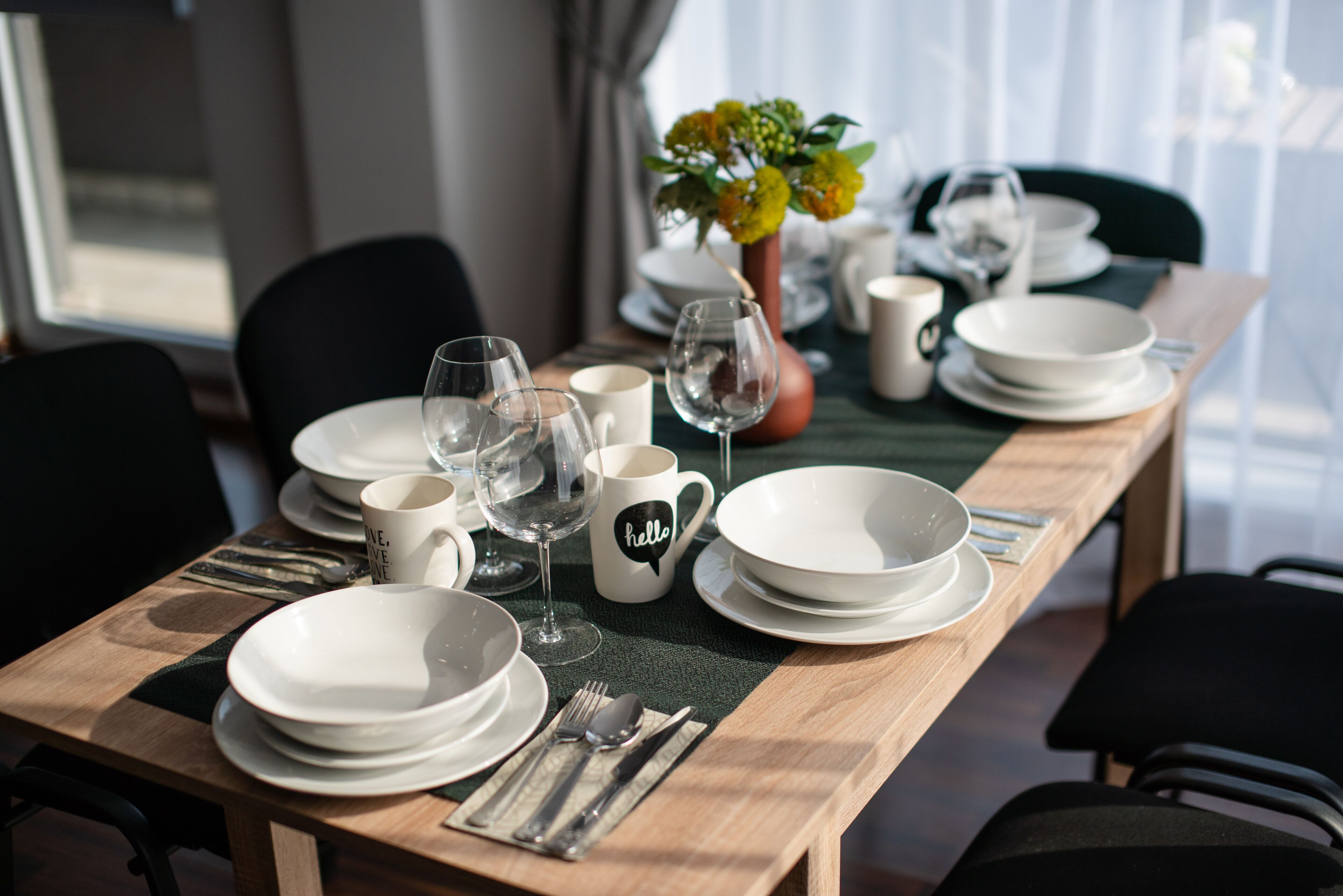 A dining table set with white plates, glasses, and cutlery, a vase with flowers, and a 'hello' mug.