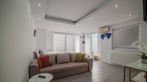 A modern living room with a beige sofa, a glass coffee table, and a window with blinds. - Niagara Apartments (Baia Mare)