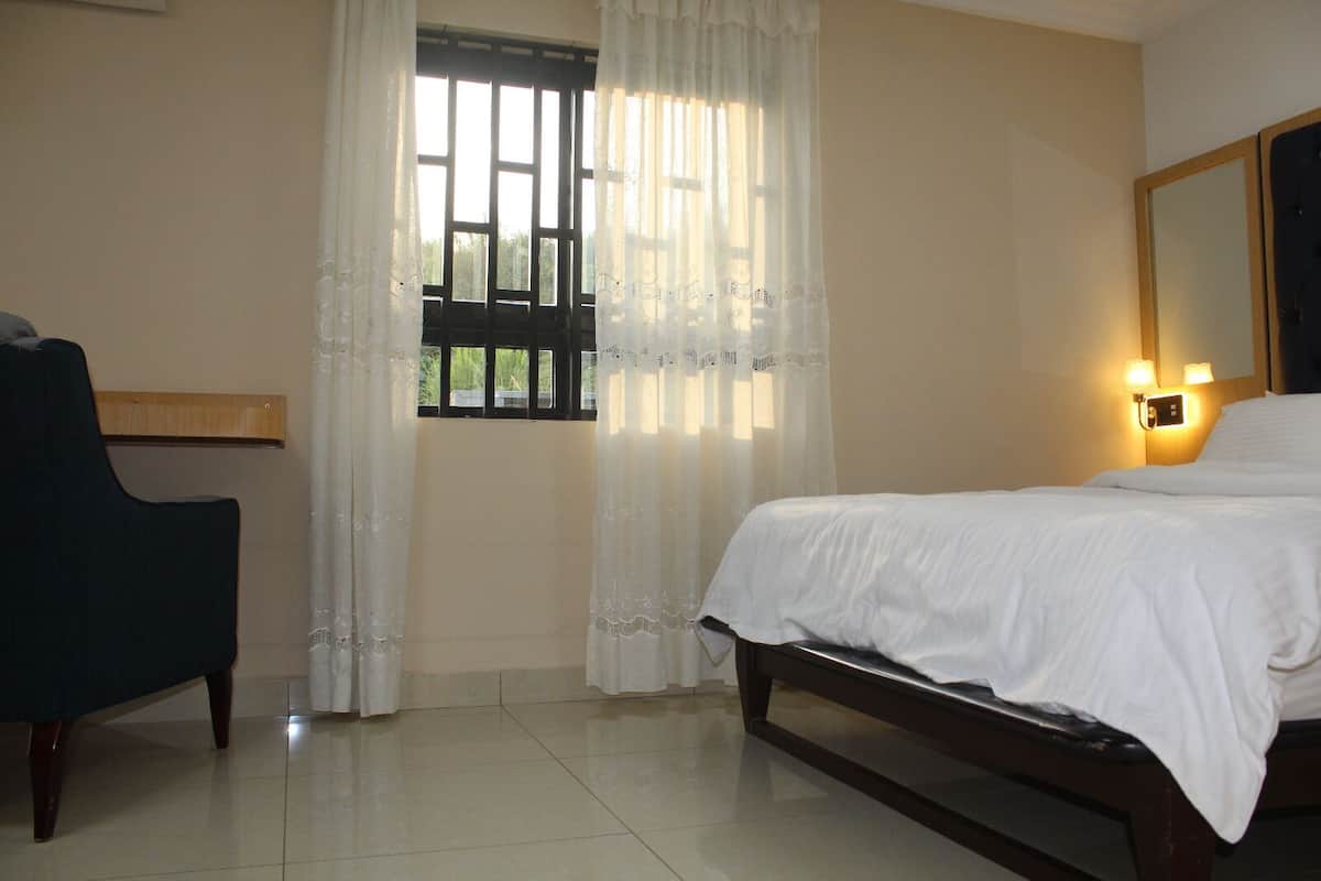 A hotel room with a bed, a chair, a window with curtains, and a wall-mounted light fixture.