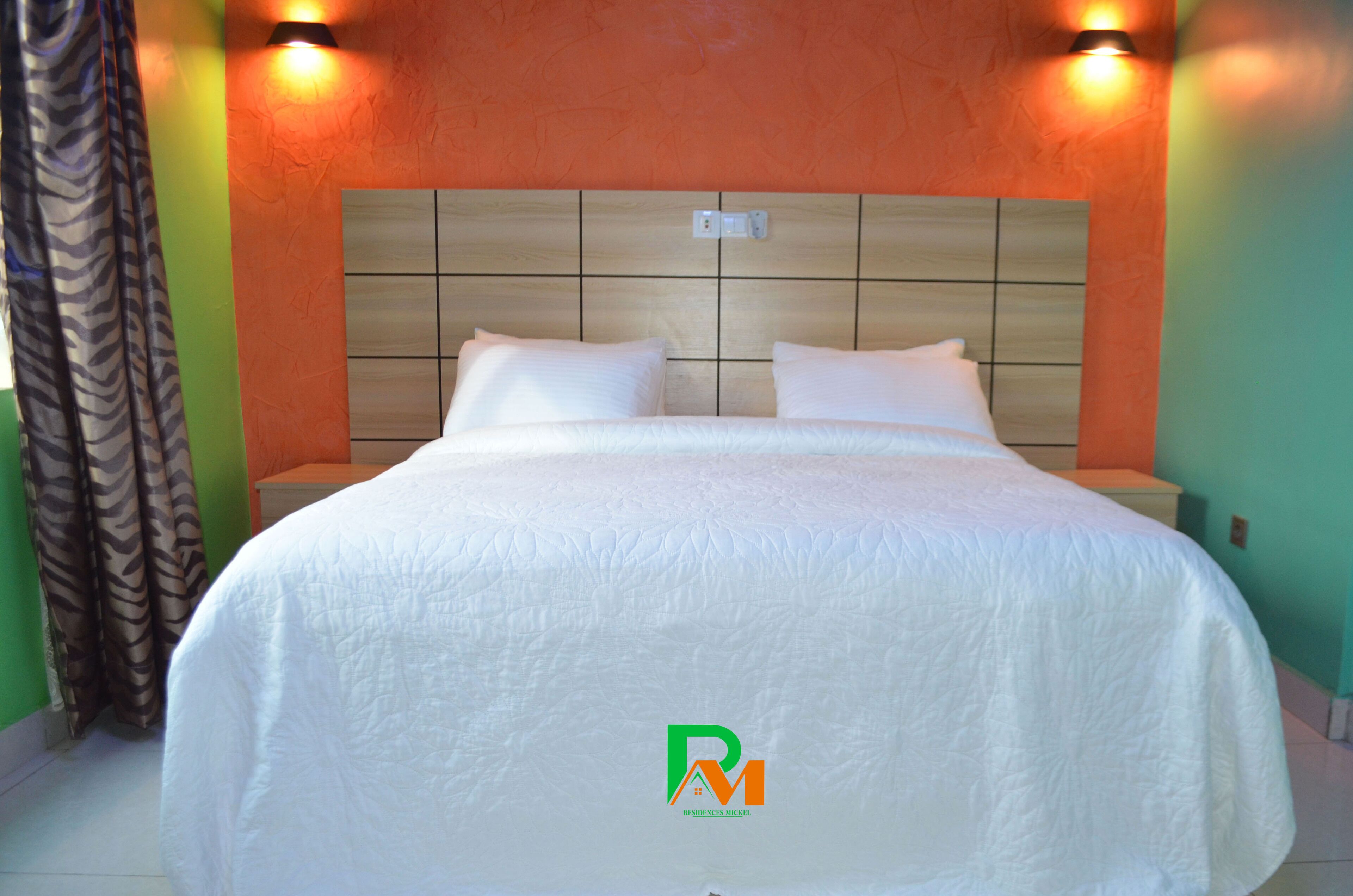 A hotel room with a large bed, two pillows, a headboard with a light switch, and a logo on the bedspread.
