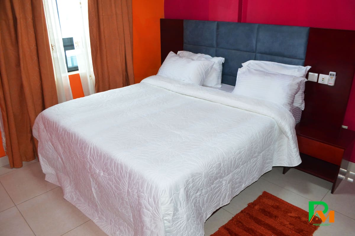 A neatly made bed with white linens in a room with a red accent wall and a brown headboard.