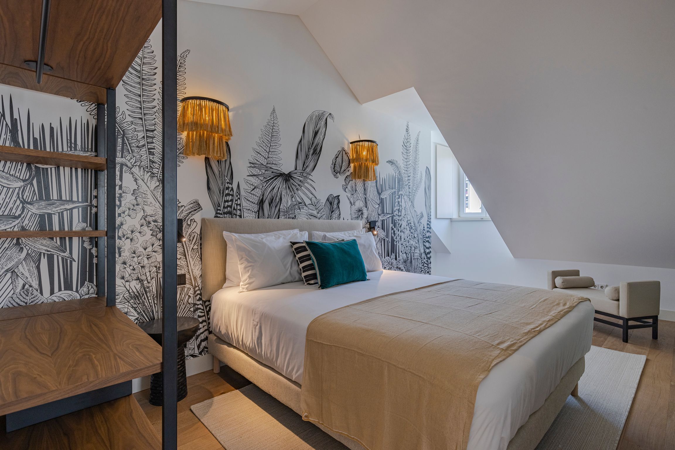 A modern bedroom with a sloped ceiling, a large bed with white bedding, a wooden side table, and a wall with black and white leaf patterns.