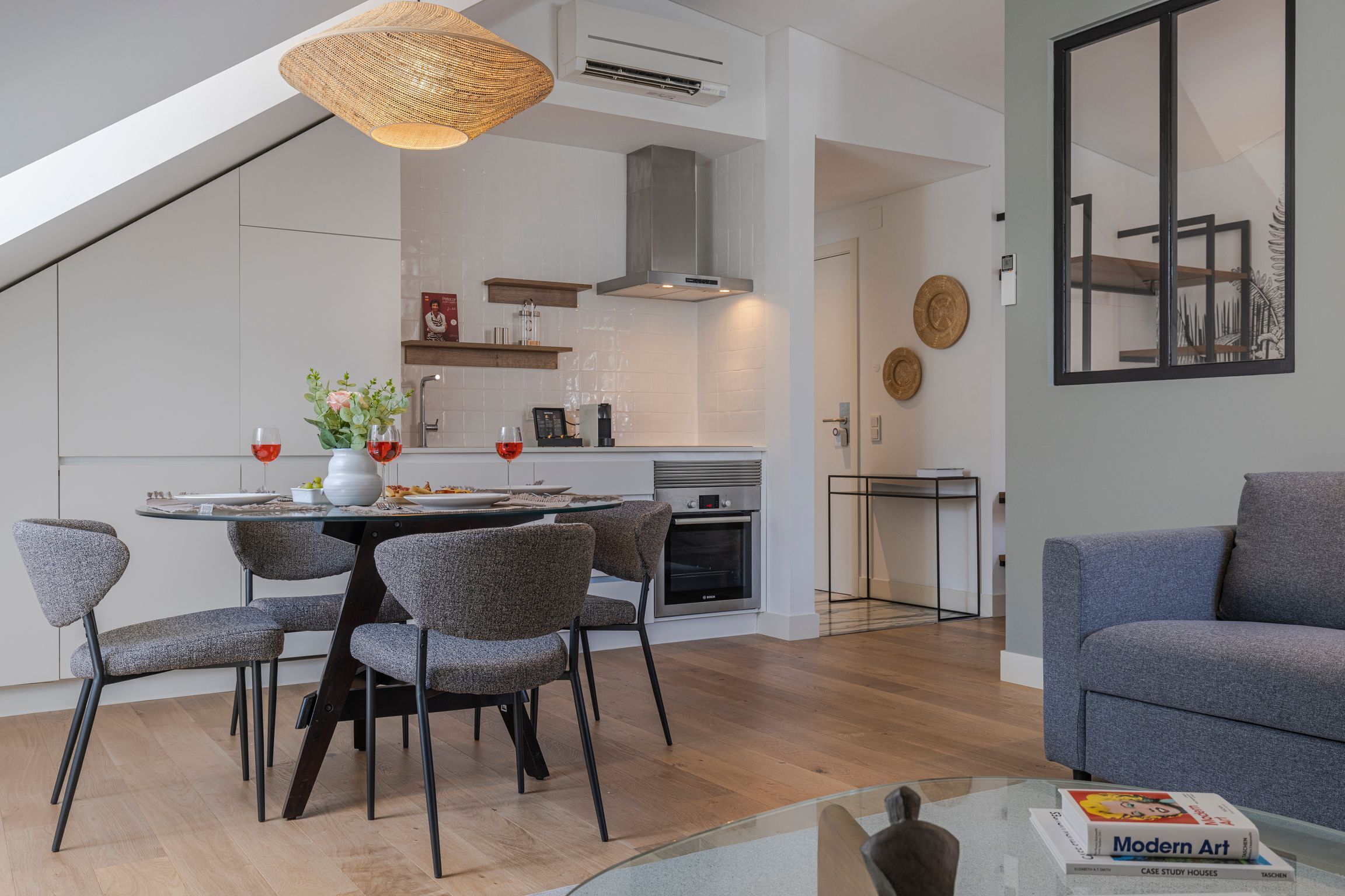 Une cuisine moderne avec des meubles blancs, un coin repas avec des chaises grises à revêtement, et un canapé gris dans le salon.
