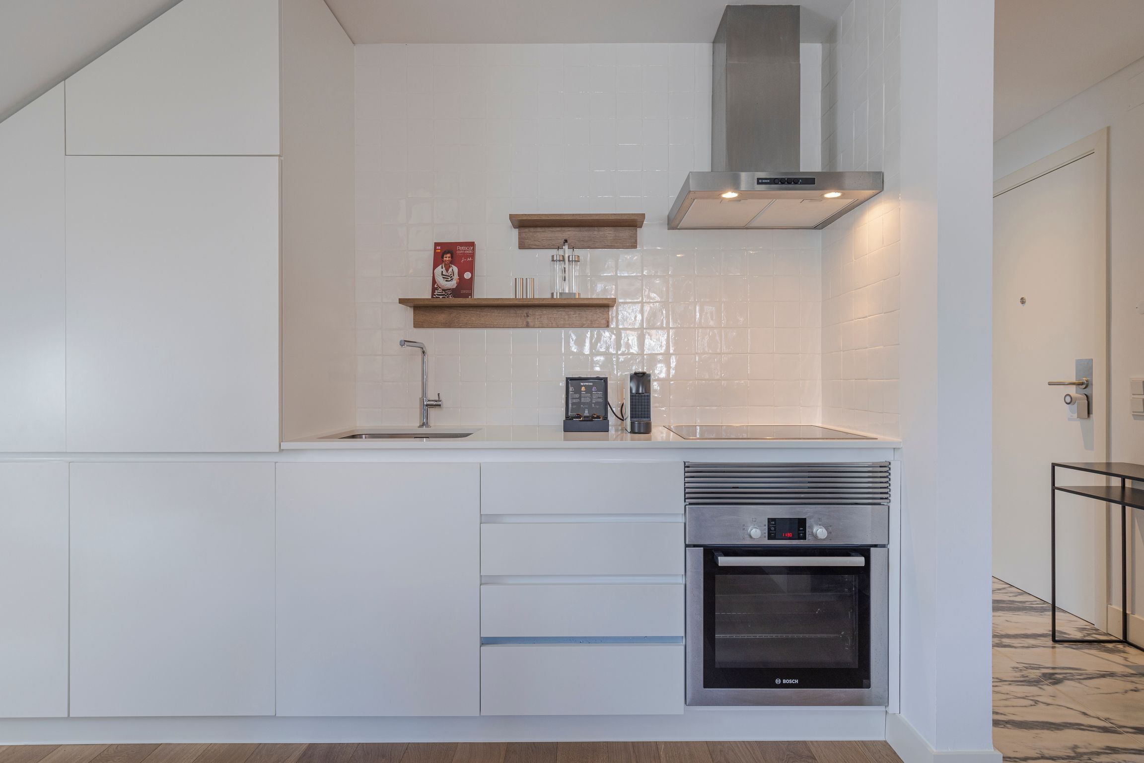 A modern kitchen with white cabinetry, a built-in oven, a stainless steel range hood, and wooden shelves.