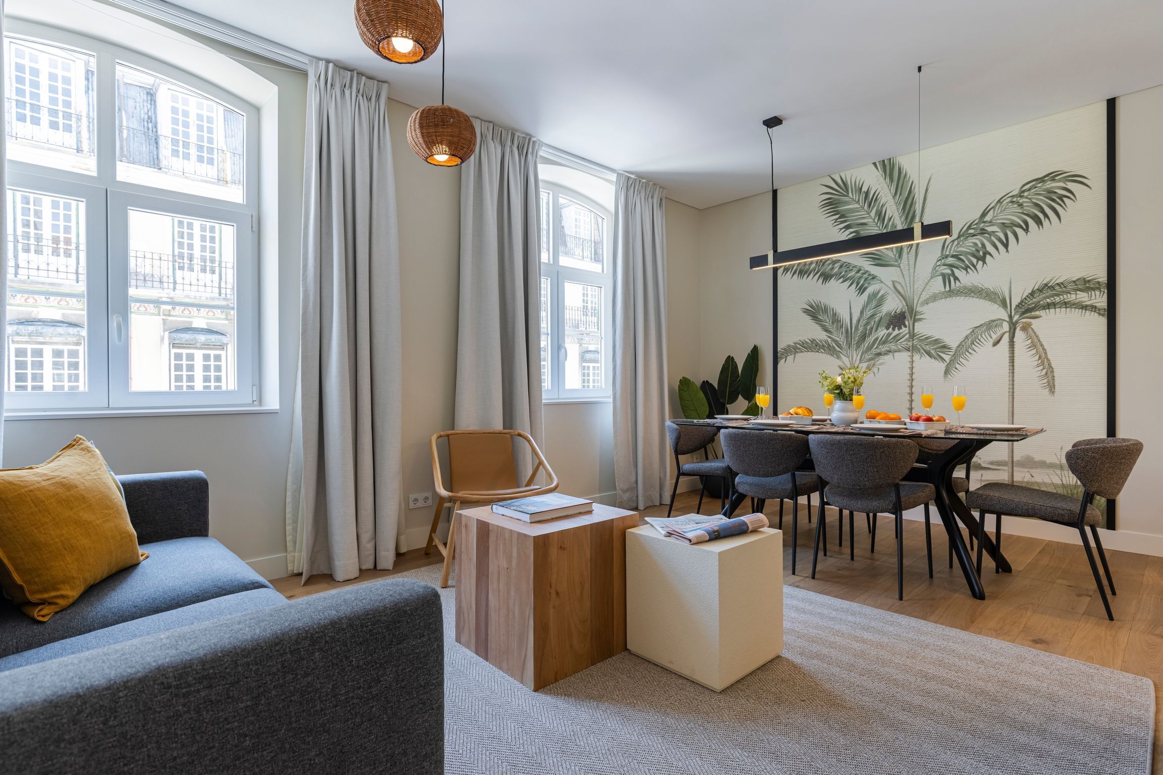 Ein modernes Wohnzimmer mit einer Couch, einem Holztisch, einem Essbereich mit Tisch und Stühlen sowie großen Fenstern mit Vorhängen.