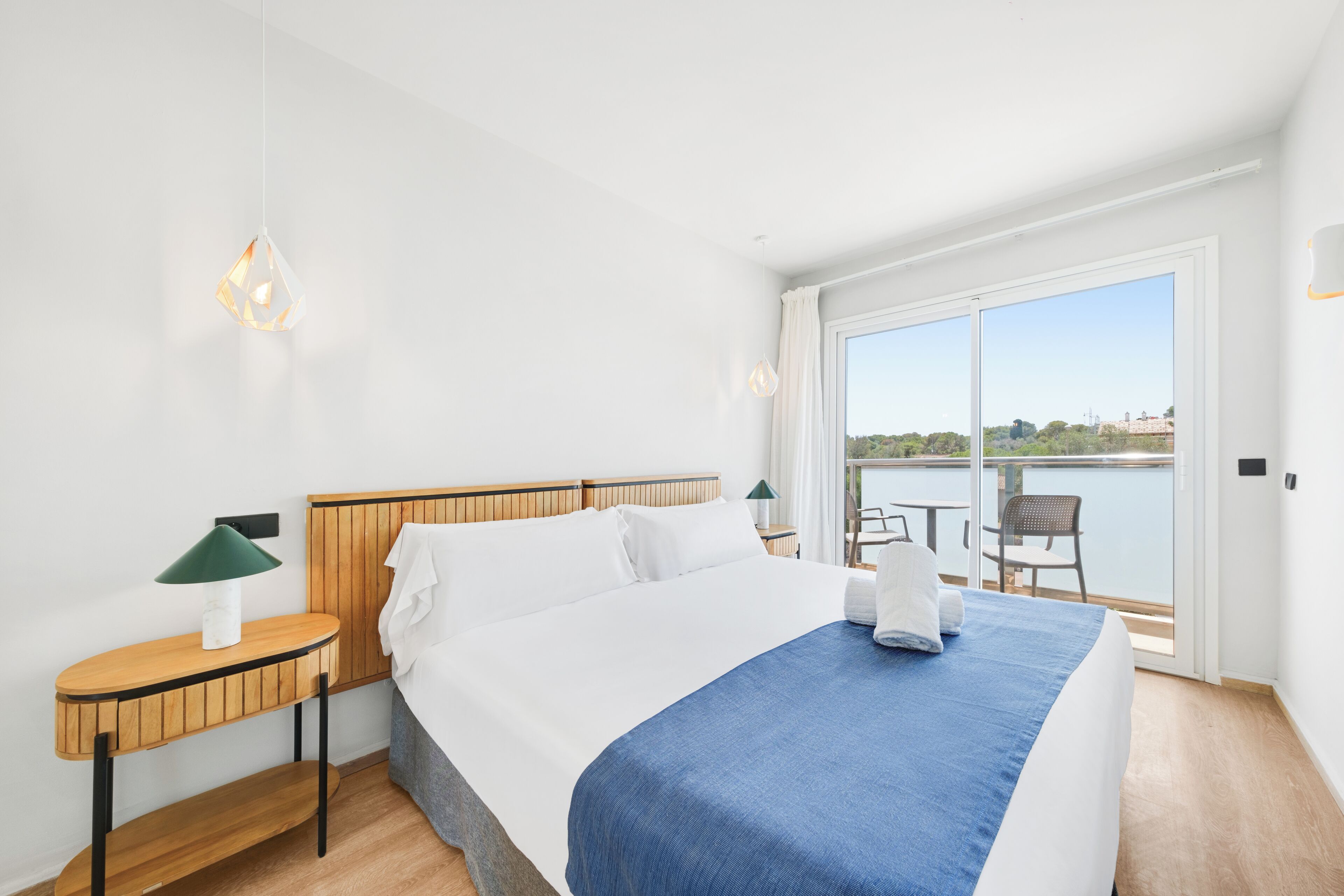 Habitación de hotel con cama, mesita de noche, lámpara, silla y vista al agua.
