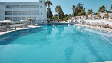 Una gran piscina con sillas de descanso y sombrillas, rodeada de palmeras y un edificio blanco con varios balcones.